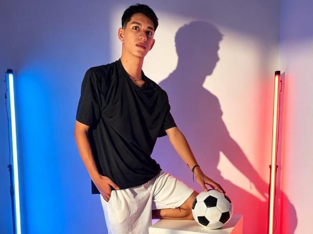 NicolasRosenthal stands with hands in white shorts, black shirt, black soccer ball on white table, blue and red lights casting shadow, short dark hair, slightly cocky, relaxed pose.