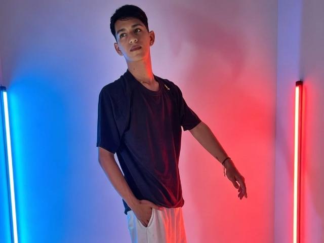 Nicolas Rosenthal, avec ses cheveux noirs courts, portant un t-shirt noir Nike et un pantalon blanc, debout, les mains dans les poches, avec des lumières néon bleues et rouges de chaque côté..