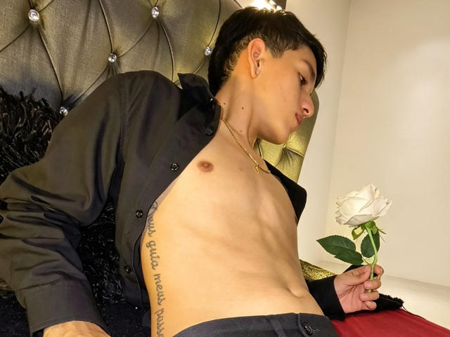 NicolasRosenthal, sem camisa, camisa preta de botões, segurando uma rosa branca, tatuagem no lado direito, colar de ouro, deitado na cama, cabeceira cinza com textura, cabelo preto curto, perfil lateral, ambiente de quarto..