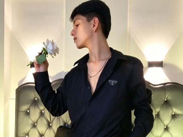 NicolasRosenthal, um homem asiático, camisa preta, cabelo preto curto, segurando uma rosa branca, perfil lateral, pele clara, pequenas brincos, cabeceira verde com textura, arandelas na parede, camisa ligeiramente aberta, colar..