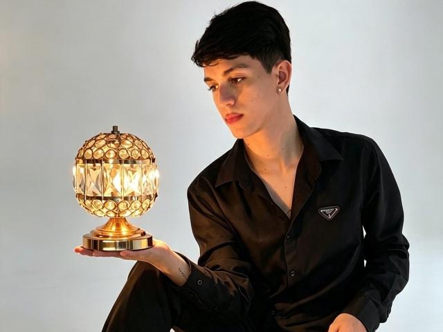 NicolasRosenthal with short black hair, black shirt, holding a golden, patterned lamp with a glowing bulb in his right hand, wearing a small earring on his left ear.