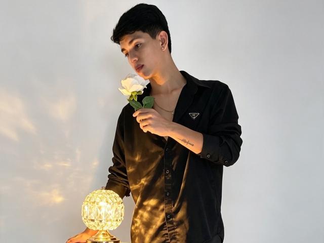 NicolasRosenthal, with short black hair, wearing a black button-up shirt, holds a white rose to his nose, and holds a glowing crystal lamp in his other hand.