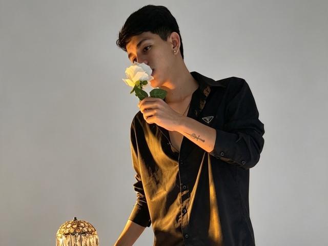 NicolasRosenthal, with short black hair, wearing a black button-up shirt, smelling a white rose, has a small cross tattoo on his right forearm, and a small earring.