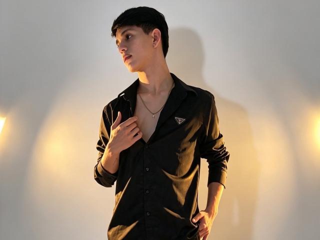 NicolasRosenthal, magro, com cabelo preto curto, vestindo uma camisa preta desabotoada, revelando um pouco do peito, segurando a camisa com a mão esquerda, com a mão na cintura, sombra na parede..