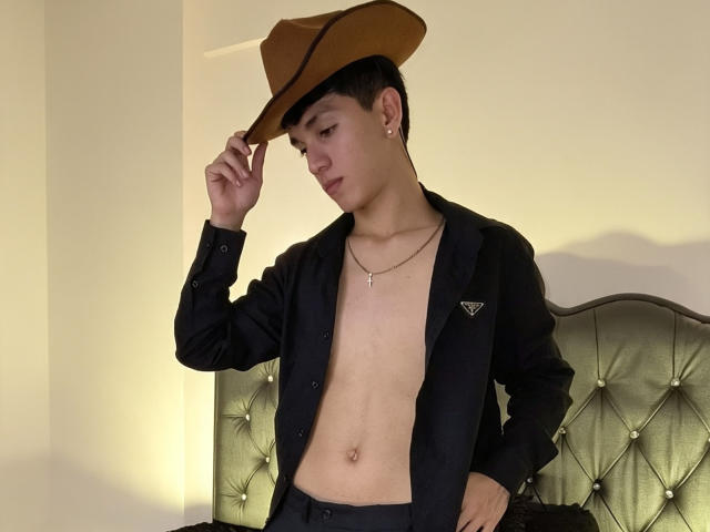 NicolasRosenthal with a brown cowboy hat, black jacket, and no shirt, exposing his flat, bare chest and belly. He has a small earring, necklace, and dark pants.