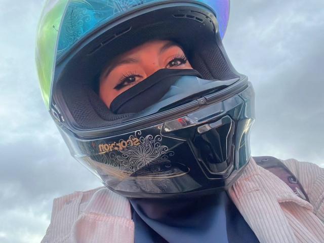 PaulaLoughty with a shiny black motorcycle helmet, blue-green visor, and black mask, wearing a pink striped shirt, her eyes looking down with long eyelashes.