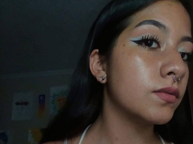PaulaLoughty has dark brown hair, thick black eyebrows, white eyeliner, and a nose ring. Her skin is tan, and she's wearing a white halter top. Her lips are pink.