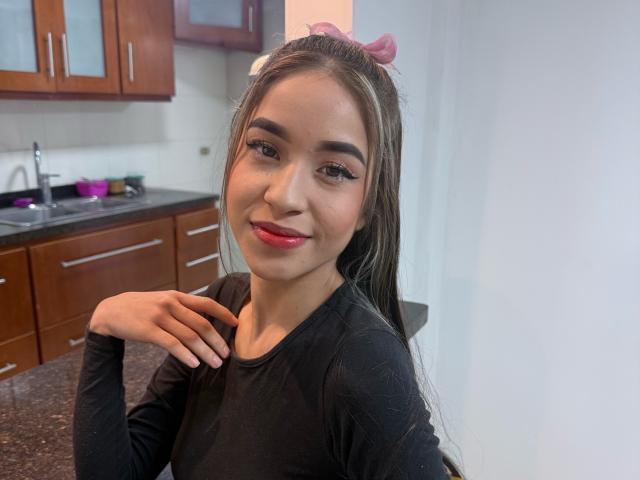 Sammantha Olssen, avec de longs cheveux bruns, un nœud rose, une chemise noire à manches longues, du rouge à lèvres et un maquillage discret, sourit à l'appareil photo, la main droite près de la clavicule, dans une cuisine..