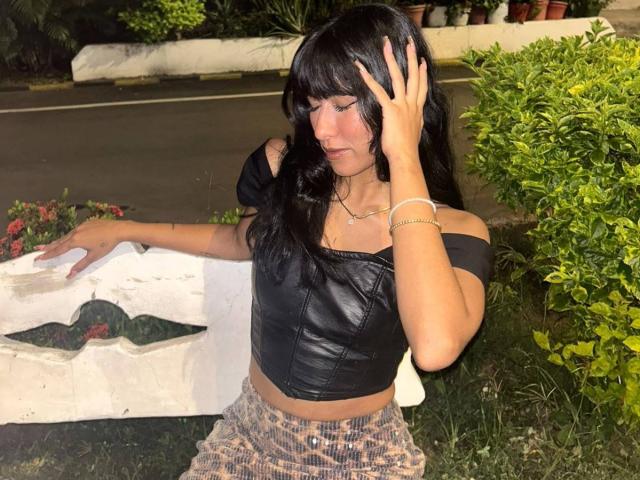 DariaShark con una blusa de cuero negro sin mangas, cabello negro ondulado y pantalones a cuadros, sosteniéndose la cabeza con una mano, de pie junto a un paño blanco y un arbusto verde..
