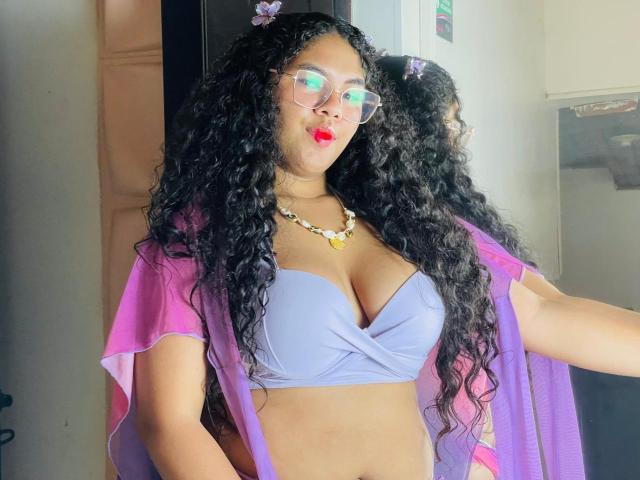 AshleyJenner with long curly black hair, wearing glasses, pink transparent robe, purple bra, necklace, and red lipstick, showing her big tits and tan skin. Reflection in mirror.