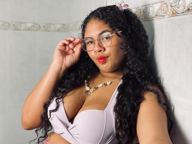 AshleyJenner with curly black hair, glasses, red lipstick, and a lavender bra, showing her tits, wearing a shell necklace, against a tiled wall.