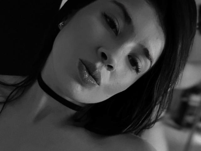 EmmaLombardi's face in black-and-white, with her head tilted, wearing a choker, and her glossy lips slightly parted, looking seductively at the camera.