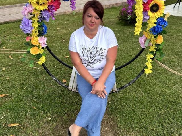 CandyFerdinand hat einen wohlgeformten Hintern und sitzt auf einer schwarzen Schaukel mit bunten Blumen, trägt ein weißes Hemd, blaue Hosen und ein rotes Armband, und darunter ist grünes Gras..
