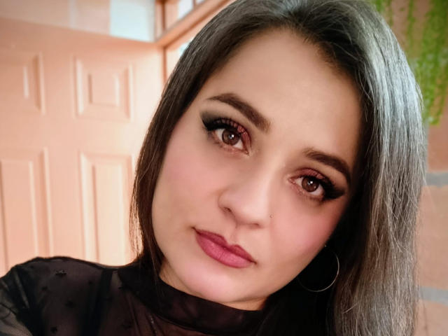 EmmaSant tiene ojos marrón oscuro, pestañas negras y gruesas, lápiz labial rosa y aros de aro negros. Lleva una blusa negra transparente, y su cabello negro lacio le cae de un lado..