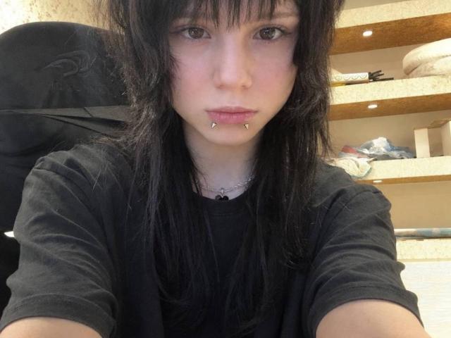 Micaela Legler, mit schwarzen Haaren, heller Haut und einem gestochenen Unterlippenpiercing, trägt ein schwarzes Hemd und hat ein Selfie gemacht. Ihre kleinen Brüste sind teilweise sichtbar. Sie sitzt auf einem Stuhl..