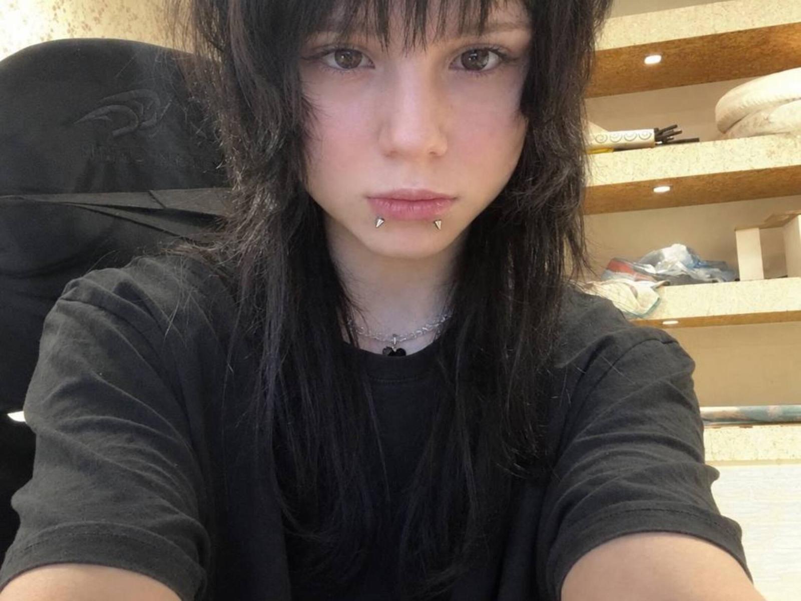 MicaelaLegler with black hair, pale skin, and pierced lower lip, wearing a black shirt, in a selfie. Her small tits are partially visible. She's in a chair.