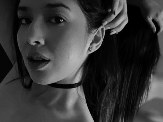 Black-and-white photo of EmmaLombardi with her hand in her long, dark hair, wearing a black choker, slightly parted lips, and a small earring in her right ear.