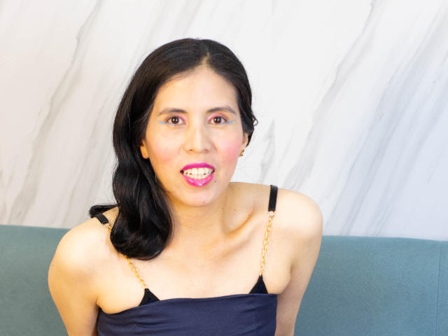 LarisaBrown with black hair, pink lipstick, and a chain strap dress, showing a toothy grimace and a small drop of saliva on her chin. Her tits are visible.