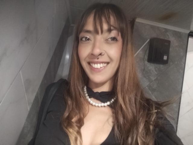 IsaChaud with brown hair, bangs, nose ring, smiling, wearing black top, pearl necklace, showing cleavage, standing in tiled bathroom, visible black hand dryer in background.