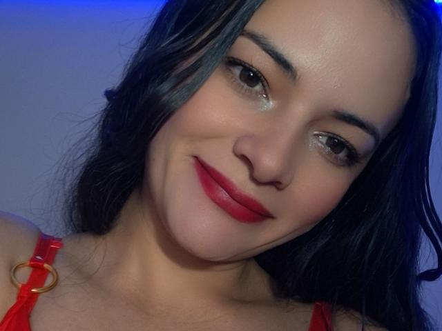 EstheraBloom, met glanzende bruine ogen, rode lippenstift en zwart haar, draagt een rode top met een gouden ring en glimlacht zachtjes. Haar huid is licht en ze is schattig..