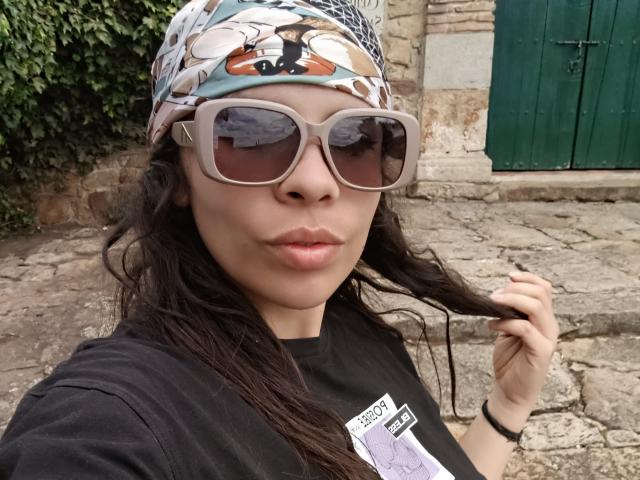 VioletaStone with long black hair, wearing a patterned headscarf, large gray sunglasses, black t-shirt, and holding her hair, standing on a cobblestone street.