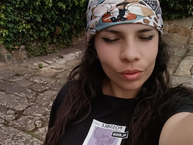 VioletaStone has a cute face with light skin, dark wavy hair, and a patterned headscarf, wearing a black shirt with a graphic, and her eyelashes are long and pointed.