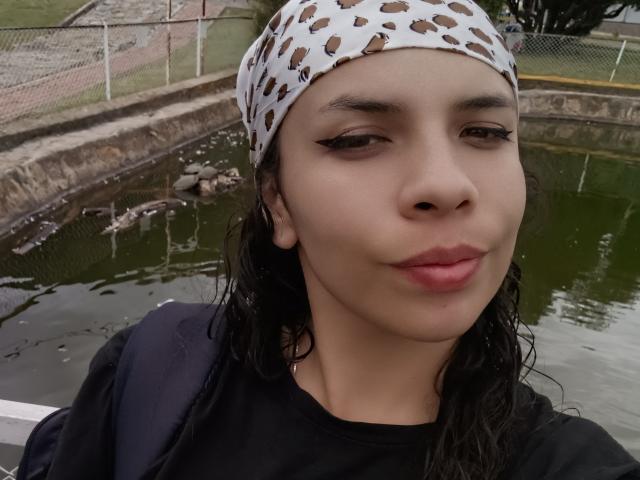 VioletaStone with wet black hair, wearing a leopard-print headscarf and black shirt, has small tits, light brown skin, and a slight smile, standing by a green, muddy water source.