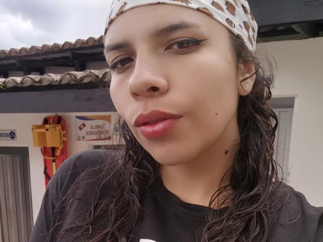 VioletaStone has wet dark hair, a white crochet headband, and wears a black shirt with red lipstick, showing a small beauty mark on her right cheek.