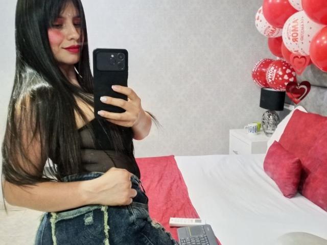 Madisson Fontaine, con cabello negro largo, lápiz labial rojo, una blusa negra transparente y jeans azules, tomando un selfie en un dormitorio con globos rojos y blancos y una cama con almohadas rojas..