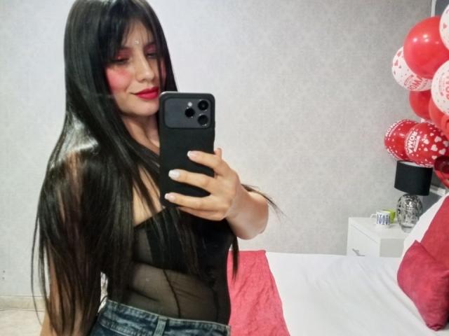 MadissonFontaine, con cabello negro largo, maquillaje rojo, una blusa negra transparente y pantalones vaqueros, está tomando una selfie frente a un espejo, mostrando sus senos. En el fondo, hay globos rojos y blancos..