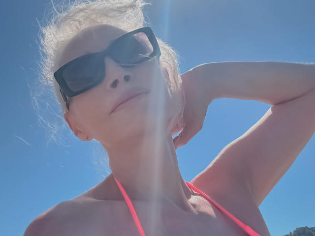 Rita Farell, con i capelli biondi, indossa occhiali da sole neri, un top rosa a fascia e ha dei riflessi del sole sul viso, mentre alza il braccio destro, sotto un cielo azzurro limpido. Il suo seno è leggermente visibile..