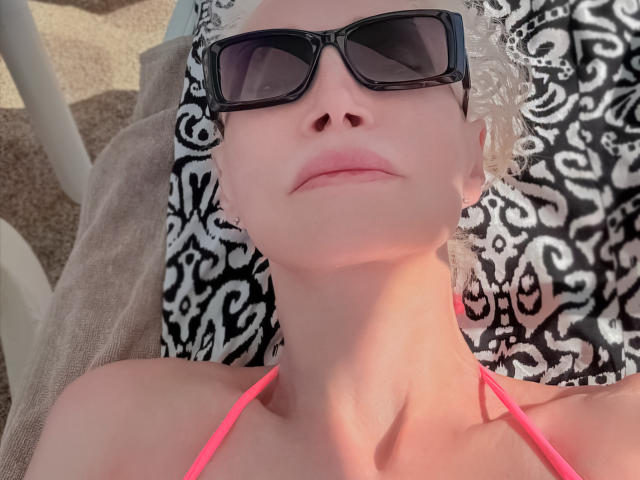 RitaFarell, com pele clara, cabelo curto e cacheado, branco, óculos de sol pretos, top rosa de alças, deitada, iluminada pelo sol, toalha preta e branca com padrão, relaxada, sem maquiagem, sorriso leve..