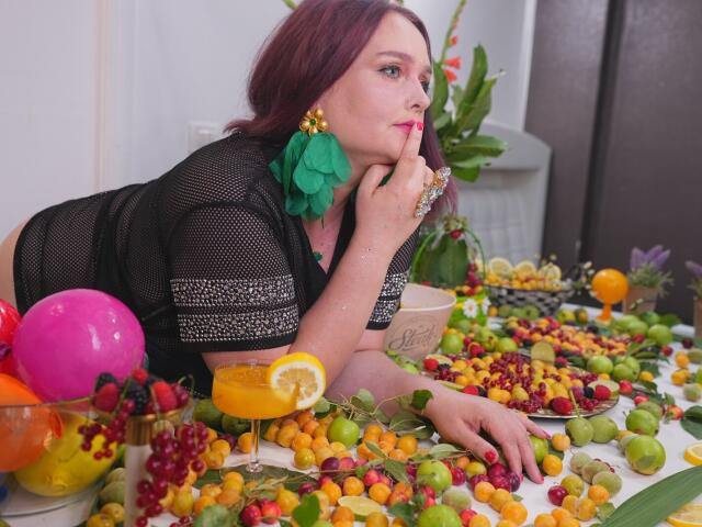 SinFulTitsDD with red hair, black mesh top, green flower earring, finger on chin, looking side, lying on fruit table with grapes, oranges, lemon, and colorful balloons.