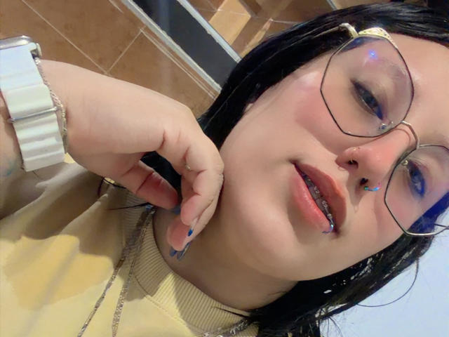 NatalieHarris with blue eyeshadow, glasses, nose ring, lip piercing, and blue nail polish, wearing a yellow sweater, touching her chin with a white watch on her wrist.