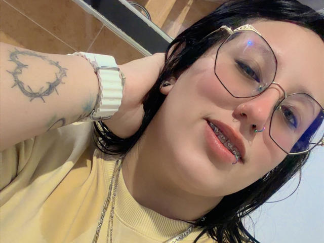 NatalieHarris with glasses, nose ring, and septum piercing, wearing a yellow shirt, showing a crown tattoo on her arm, white watch, and a silver necklace, smiling.
