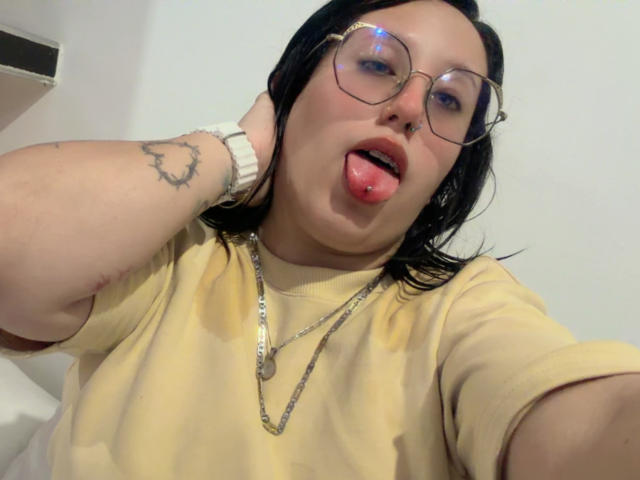 Natalie Harris met zwart haar, een bril, een geel shirt, haar tong uitgestoken, een neusring, een tatoeage op haar linkerarm, een witte horloge, een ketting en een licht geopende mond. Ze steekt haar tong uit..