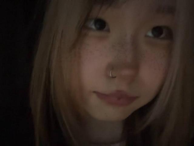 JungMcgranor has a nose ring, freckles, and light brown, straight hair with bangs. She has a serious, slightly pouty expression with dark eyes. Her face is in shadow.