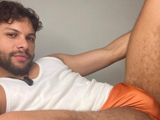 Dmalessandro with curly brown hair, beard, and white tank top, wearing orange underwear, lying down with a leg raised, showing his hairy balls and a slight smile.