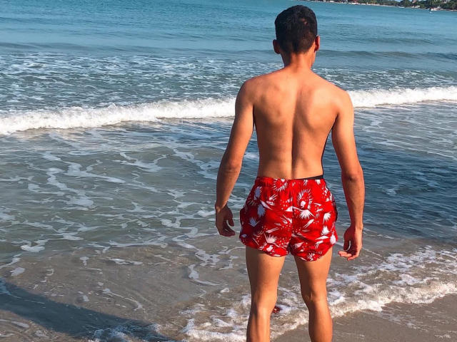 Dmalessandro, torse nu, avec un dos bronzé et musclé, et vêtu de shorts de bain à fleurs rouges, se tient dans une eau peu profonde et mousseuse, face à l'horizon. Ses fesses sont légèrement visibles..