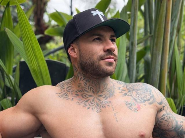 Wkite, ohne Hemd, mit Tattoos, die seine Brust und seinen Hals bedecken, trägt eine schwarze Baseballkappe, hat einen muskulösen Körperbau, einen Bart und blickt mit einem leichten Grinsen zur Seite..