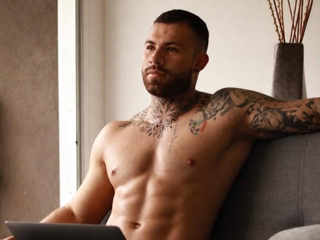 Muscular Wkite with tattoos, short brown hair, and beard, sitting shirtless on gray couch, holding laptop, looking serious, chest and arm tattoos, right arm resting on back.