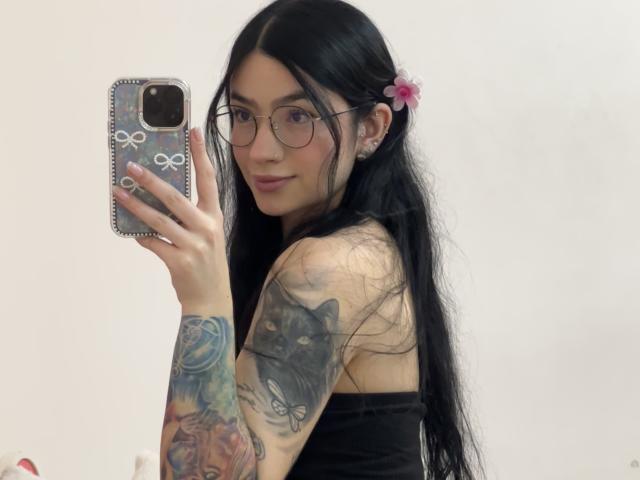 EvaaFarix with long black hair, glasses, pink flower, tattooed arm, black strap top, holding phone, taking mirror selfie, showing black cat tattoo on upper arm.