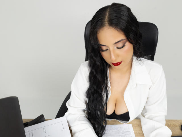 StefanyDelly with long black hair, red lipstick, and cleavage in a white shirt, sitting at a desk, looking down at a laptop and papers.