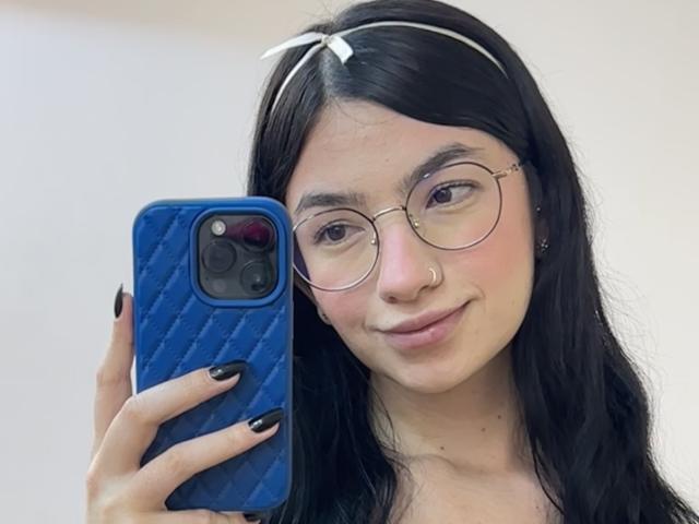 EvaaFarix with black hair, glasses, nose ring, and white headband, holding blue phone with black nails, smiling, showing cute face. Pussy not visible.