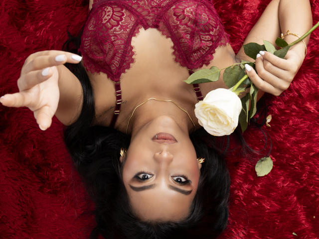 EmmaEstreilla's tits in red lace, lying on red shag, holding white rose, her face upside-down, looking at camera, with a gold necklace and white nails.