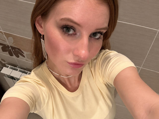 Ardelle Cooksey, una mujer pelirroja con ojos verdes, vestida con una camisa amarilla, aretes de diamantes y un collar de cadena, tomando un selfie, mostrando un rostro ligeramente sonrojado y labios rosados..