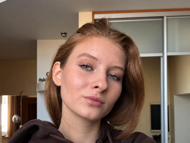 ArdelleCooksey has fair skin, green eyes, and pink lips with a slight pout. She's wearing a brown hoodie, with brown, shoulder-length hair. She's looking directly at the camera.