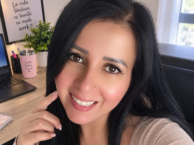Agatha Brunner, con su largo cabello negro y sus ojos marrones, sonríe a la cámara, mostrando sus labios rosados y brillantes y una pequeña marca de nacimiento en su mejilla derecha. Su mano toca su barbilla..