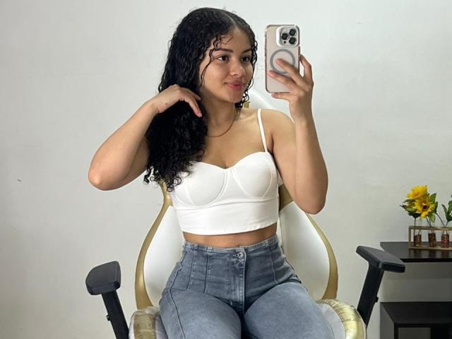 CurlyLorene mit lockigem, schwarzem Haar, einem weißen BH, der ihre Brüste zeigt, und blauen Jeans, die ein Selfie mit einem goldenen Stuhl macht, ein Handy in der Hand hält, lächelt und ein kleiner Tisch in der Nähe steht..
