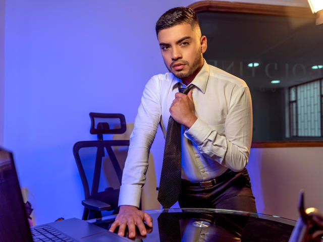 AlexanderDiaz, with a serious face, adjusting his tie, sits at a desk, wearing a white shirt and black tie, hand on a laptop, in a blue-lit office.